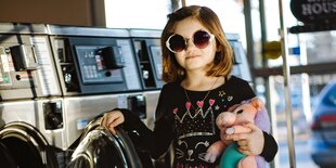 Ein Mädchen mit großer Sonnenbrille und Kuscheltier in der Hand steht an einer Waschmaschine