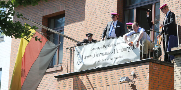 Herren in komischer Kleidung stehen auf einem Balkon und präsentieren die aktuelle Deutschland-Fahne