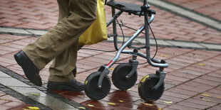 Die Beine eines Mannes sind zu sehen, der auf einer nassen Straße mit einem Rollator läuft