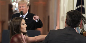 Donald Trump hält eine Rede an einem Rednerpult und zeigt mit dem Finger auf einen Mann vor ihm. Dem Mann wird von einer Frau sein Mikrofon weggenommen
