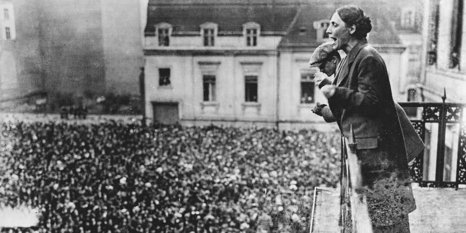 Schwarz-weiß-Foto einer Frau, die von einem Balkon zu einer Menschenmenge auf einem Platz spricht