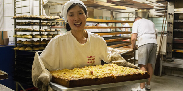 Eine Frau in Bäckerkleidung hält ein Blech Streuselkuchen in beiden Armen