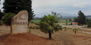 Ein Stein mit der Aufschrift "Villa Baviera" steht vor einer Palme, im Hintergrund die Bergkette der Anden