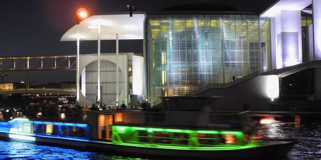Ein beleuchtets Bundestagsgebäude bei Nacht und ein bunt beleuchtets kleines Schiff davor