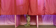 Mit rotem Stoff verhangene Wahlkabinen, in einer sind die Beine eines Mannes in Tarnhose zu sehen
