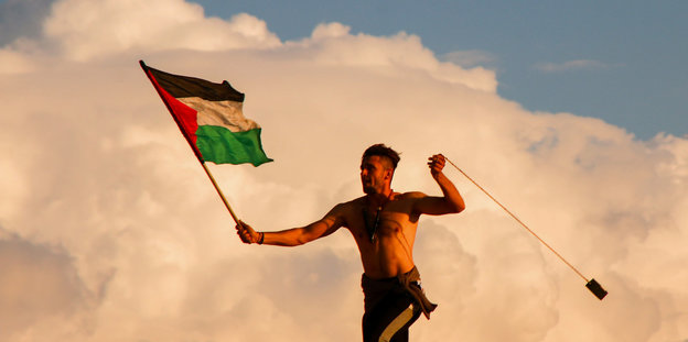 Ein Mann mit palästinensischer Fahne und einer Steinschleuder vor einer Rauchwolke