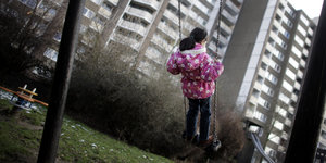 Ein Mädchen in pinker Jacke Schaukelt vor einer Kulisse aus Plattenbauten.
