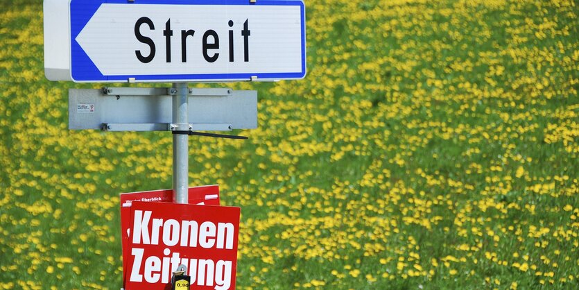 Auf einer Blumenwiese ist ein Zeitungsständer der Kronen Zeitung zu sehen, darüber ein Ortsschild nach Streit