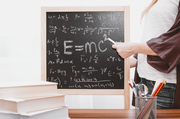 Eine Person mit einem Kreidestück in der Hand steht neben einer Tafel, die mit Formeln beschrieben ist