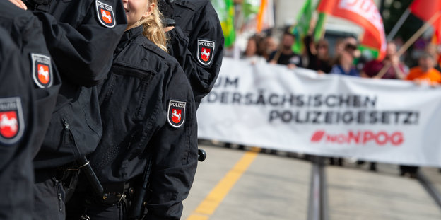 Im Vordergrund stehen niedersächsische PolizistInnen, im Hintergrund demonstrieren BürgerInnen gegen das neue Polizeigesetz