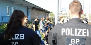 Zwei Polizisten von hinten, dahinter lehnen Menschen an einem Zaun, ihre Gesichter sind unkenntlich gemacht.