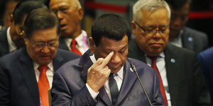 Der philippinische Präsident Rodrigo Duterte hält seinen Mittelfinger an sein Auge und sitzt zwischen zwei Personen