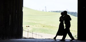 Silhouette eines tanzenden Paares in einer Art Schuppen, im Hintergrund sind grüne Hügel zu sehen