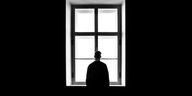 Eine Person steht einsam vor einem Fenster. Den Rücken hat sie dem Betrachter zugewandt