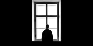 Eine Person steht einsam vor einem Fenster. Den Rücken hat sie dem Betrachter zugewandt