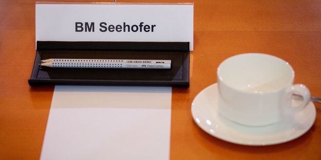 Auf einem Namenschild steht BM Seehofer. Davor liegen ein weißes DIN A4 Blatt sowie zwei Bleistifte. Daneben steht eine weiße Porzellantasse mit Untersetzer
