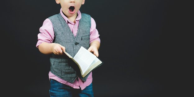 Ein kleiner Junge in einer schicken Weste hält ein aufgeschlagenes Notizbuch vor sich