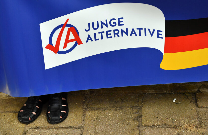 Schwarze Herrensandalen schauen unter einem Plakat der Jungen Alternativen hervor