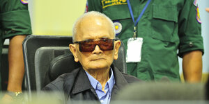 Ein alter Mann mit Sonnenbrille sitzt zwischen zwei Uniformierten