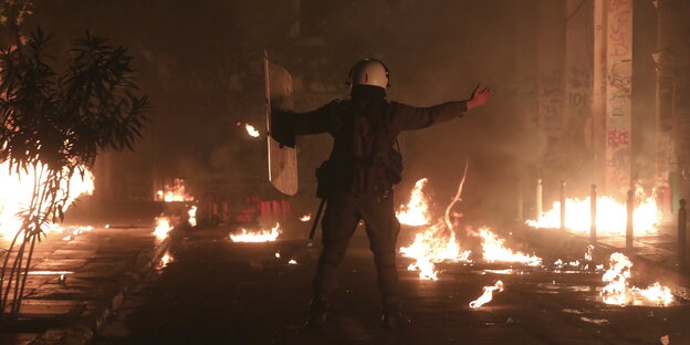 ein Polizist steht mit ausgebreiteten Armen und einem Schild in der Nacht auf einer Straße, auf der es an vielen Stllen brennt