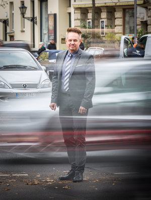 Ein Mann in Anzug steht auf einer Straße, hinter ihm fährt ein Auto vorbei