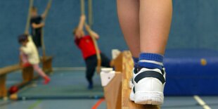 Ein Kinderbein auf einem Schwebebalken. In Entfernung Kinder, die versuchen ein Seil hochzuklettern. Seil