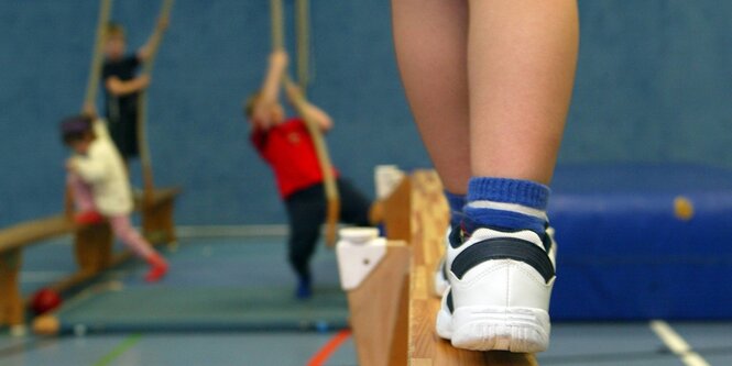 Ein Kinderbein auf einem Schwebebalken. In Entfernung Kinder, die versuchen ein Seil hochzuklettern. Seil