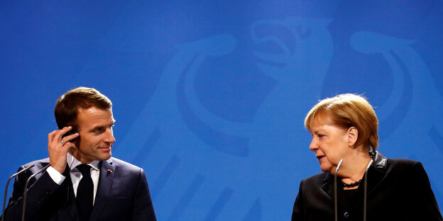 Macron und Merkel geben vor einer blauen Wand eine Presskonferenz und schauen einander an