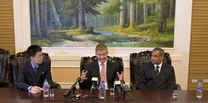drei Männer sitzen an einem Tisch, im Hintergrund ein riesiges Gemälde. In der Mitte befindet sich Erik Solheim