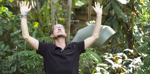 Carl von Siemens, ein schlanker Mann, ist von vielen Grünpflanzen umgeben und hebt beide Arme in die Luft