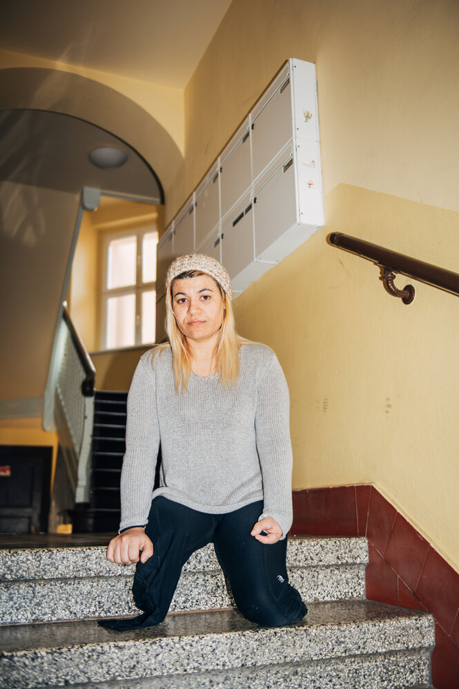 Eine blonde Frau sitzt in einem Treppenhaus, sie hat keine Beine und stützt sich auf einer Treppenstufe ab