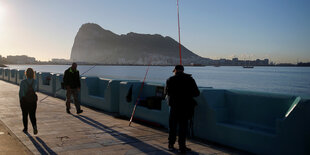 Passanten und Angler an einer Uferpromenade, im Hintergrund Gibraltar