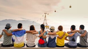 mehrere junge Leute sitzen am Wasser und haben jeweils die Arme um die Schultern ihrer Nachbarn gelegt. Man sieht sie von hinten