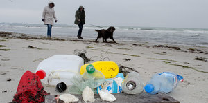 an einem Sandstrand liegen Plastikflaschen, dahinter sind Spaziergänger zu sehen