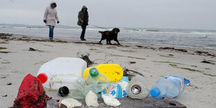 an einem Sandstrand liegen Plastikflaschen, dahinter sind Spaziergänger zu sehen