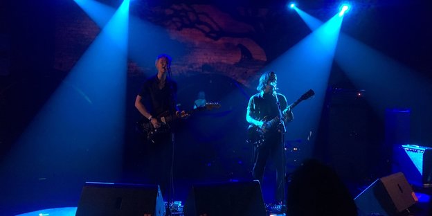 Zwei Personen mit Gitarren stehen auf einer dramatisch beleuchteten Bühne