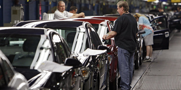 Einige Autos stehen hintereinander in einer Fabrikhalle und werden von GM-Angestellten bearbeitet