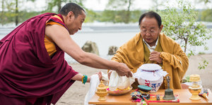 Dem buddhistischen Lehrer Rinpoche in traditionellem geleben Gewand wird von einem Mann in rotem Gewand Speisen serviert