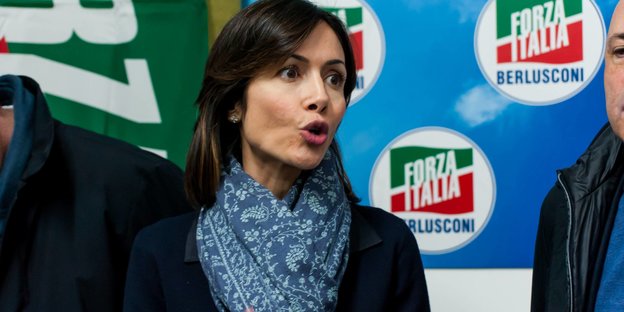 Eine brunette Frau mit blauem Mantel und Schal gestikuliert vor einem Plakat der Forza Italia