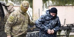ein vermummter Militär eskortiert einen Mann in blauen Tarnklamotten und in Handschellen