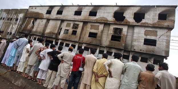 eine Reihe von Männern steht vor einer ausgebrannten Fabrik. Die Kamera hat ihre Rücken aufgenommen und wurde vom Fotografen schief gehalten