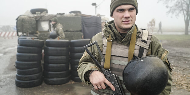 Ein Mann in militärischer Kleidung steht bewaffnet vor einem Reifenstapel, dahinter steht ein Militärfahrzeug