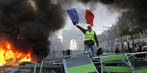 Ein Mann in einer gelben Warnweste steht neben eine brennenden Barrikade vor dem Triumphbogen in Paris