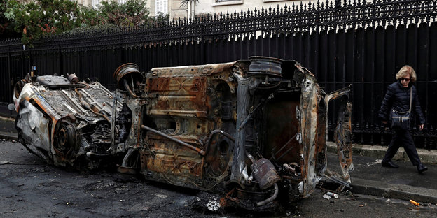 Ausgebrannte Autos auf einer Straße in Frankreich. Eine Passantin läuft mit irritiertem Blick daran vorbei