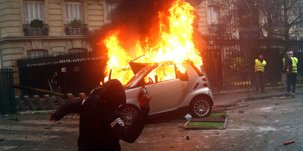 Ein vermummter Demonstrant schmeißt etwas auf ein brennendes Auto in Paris