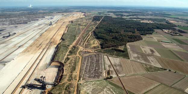 Luftbild eines Tagebaus, an dessen Rand ein Waldstück zu sehen ist