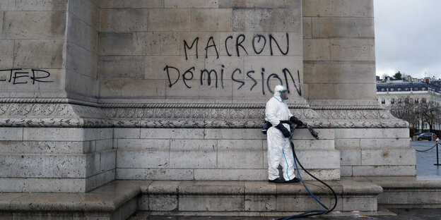 Ein Mann in weißem Schutzanzug vor einem Graffito "Macron Demission"