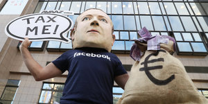 Ein Mann mit Mark-Zuckerberg-Maske hält ein Schild, auf dem "Tax me" steht