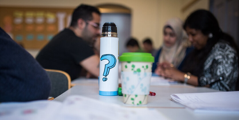 Auf einem Tisch stehen eine Thermoskanne und ein Mahrweg-Kaffeebecher. Unscharf sind im Hintergrund sitzende Menschen zu erkennen