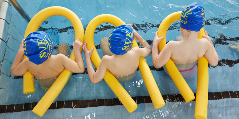 Drei Kinder sitzen am Beckenrad in Schwimmhosen und mit gelben Schwimmnudeln